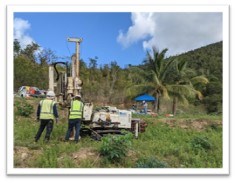 Two workers in yellow vests and hard hats stand in front of soil sampling eqipment among green vegetation and palms in the background.
