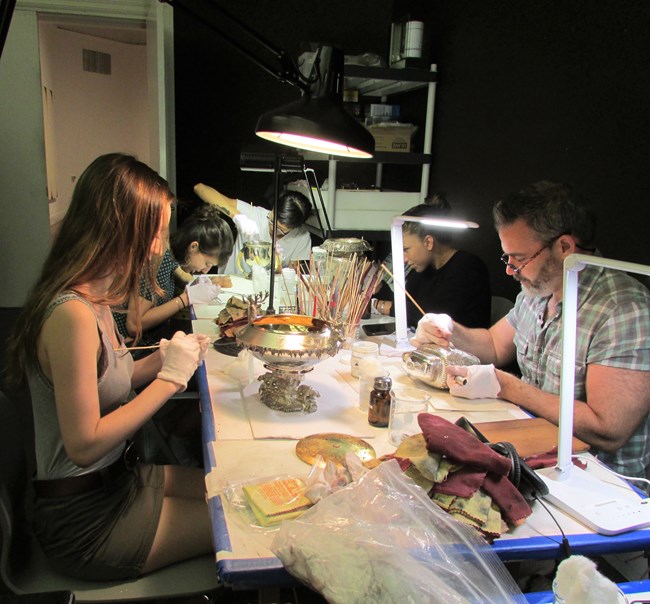 Volunteers sitting around a work table cleaning silver objects for exhibition.