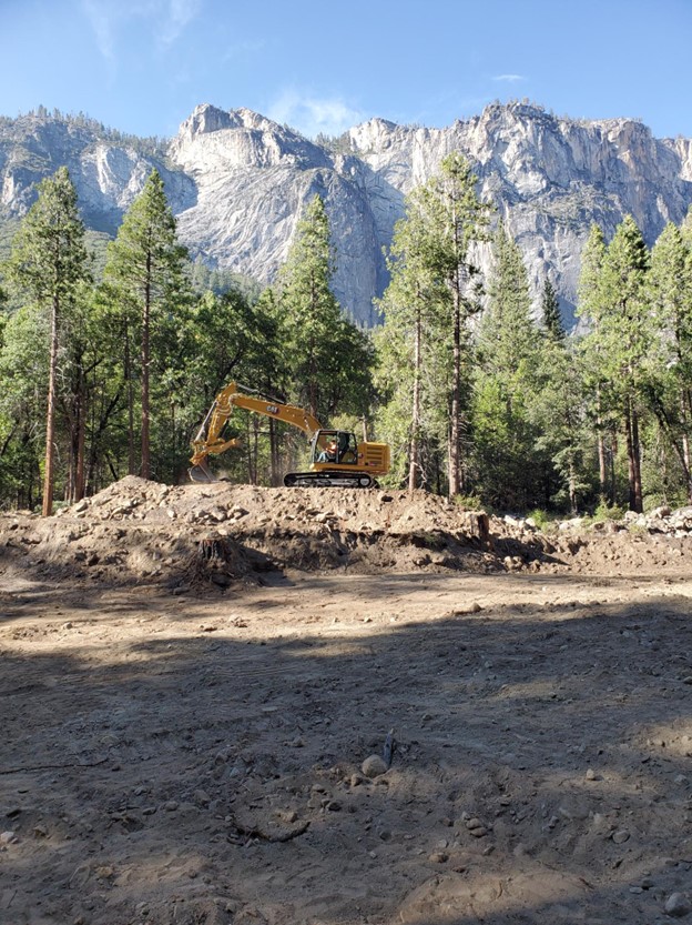 A piece of heavy equipment does "dirt work" working on soil and dirt with pine trees and granite mountains in the background.