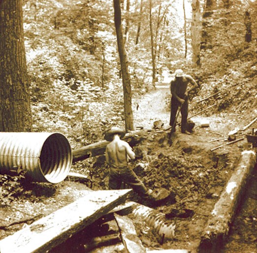 Two men dig with shovels in a wooded area next to a large pipe