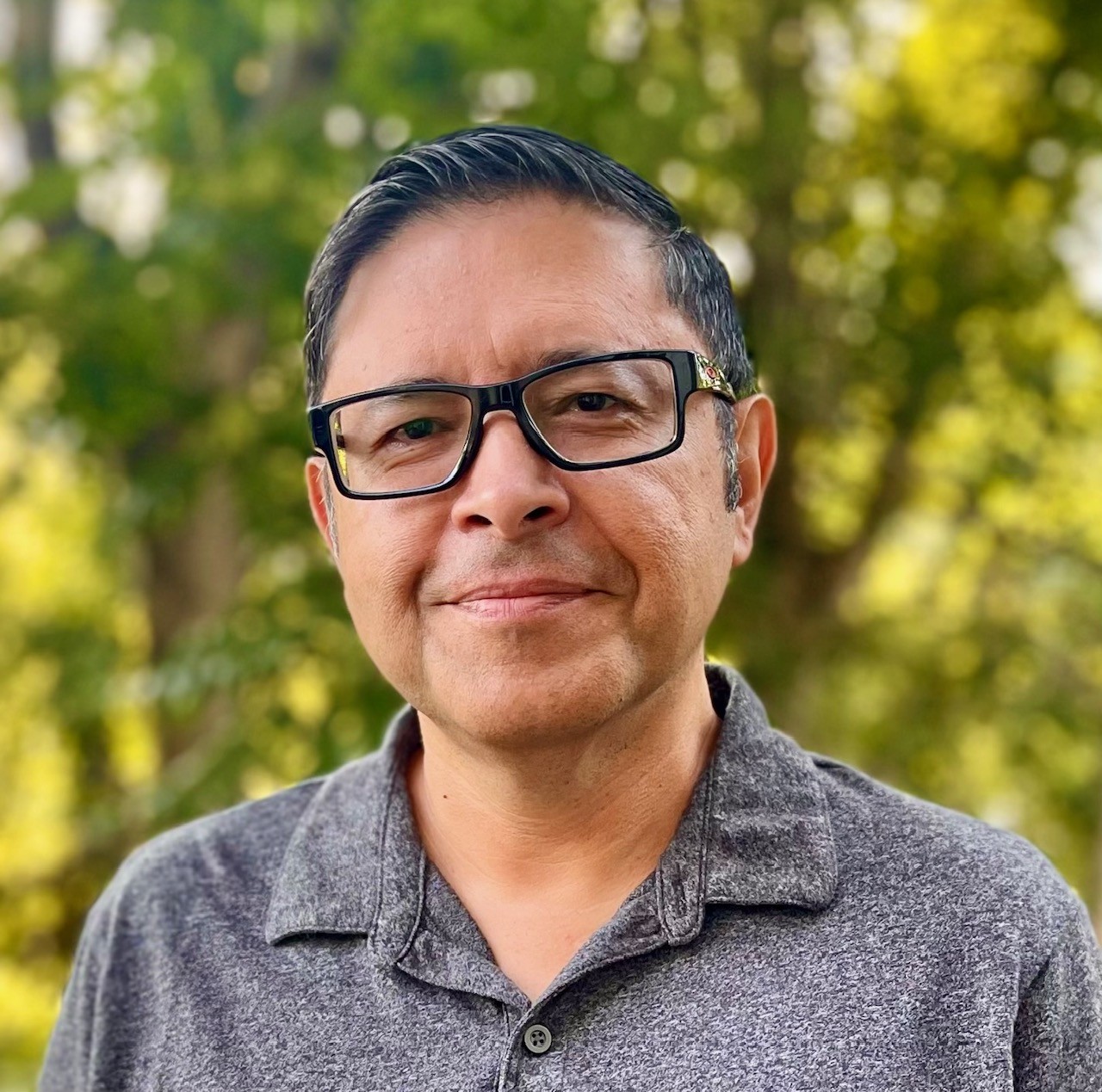 a man wearing glasses and a collared gray shirt smiles for the camera