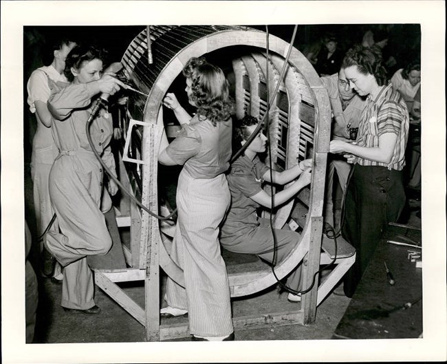 Black and white photo of five white women in pants or coveralls screwing bolts onto various parts of a cylindrical frame