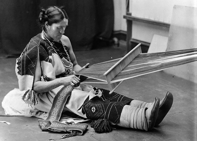 Black and white photograph of We'wha sitting with their legs outstreatched and wearing traditional Zuni attire. They are weaving with a backstrap loom.