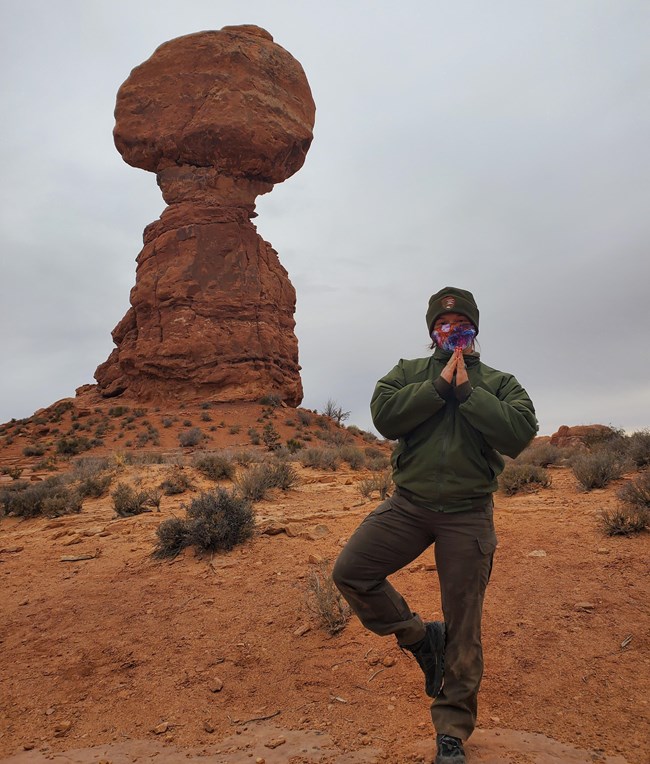 A female park ranger in uniform stands with one leg raised. Behind her is a towering orange colored rock balanced on a rock pedestal.