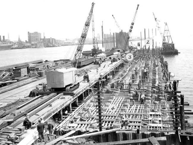 Photograph of workers and cranes on a pier in the midst of construction extending into Boston Harbor. Pile driving equipment is on a floating lighter at the edge of the pier.