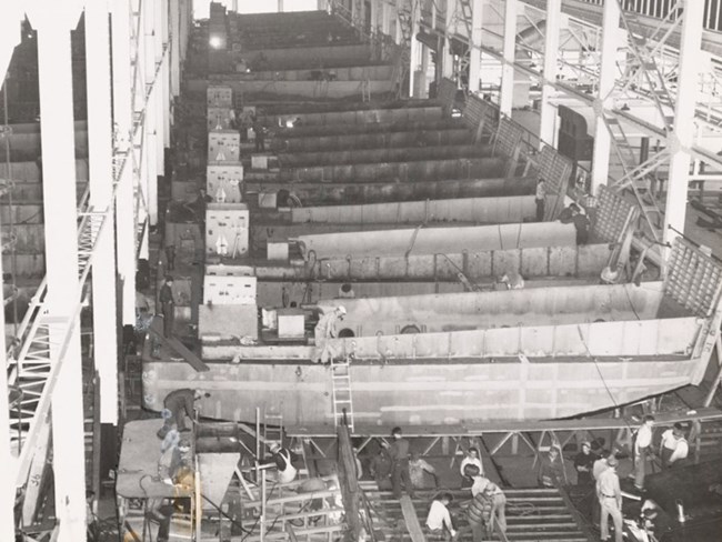 Photograph from balcony of large manufacturing structure. Workers assemble a tank landing craft in foreground. In background is a long line of assembled landing craft.