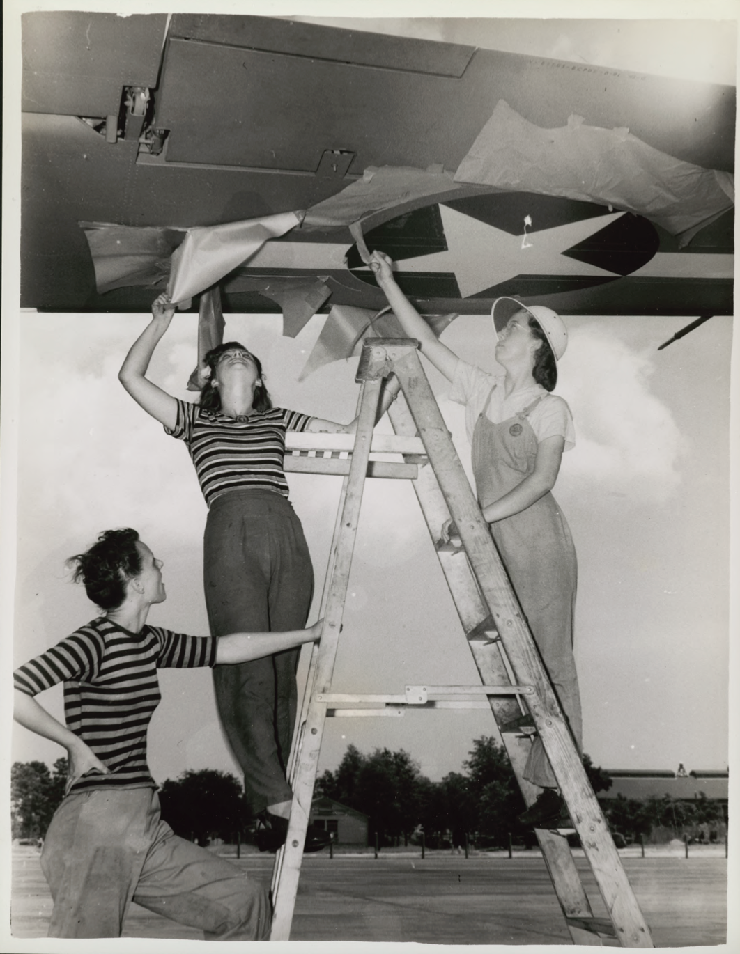 black and white photo of three white women, two standing on a ladder, working on a ceiling