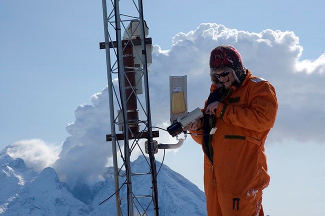 A scientist works with instruments on a mountain top with an active volcano behind him.