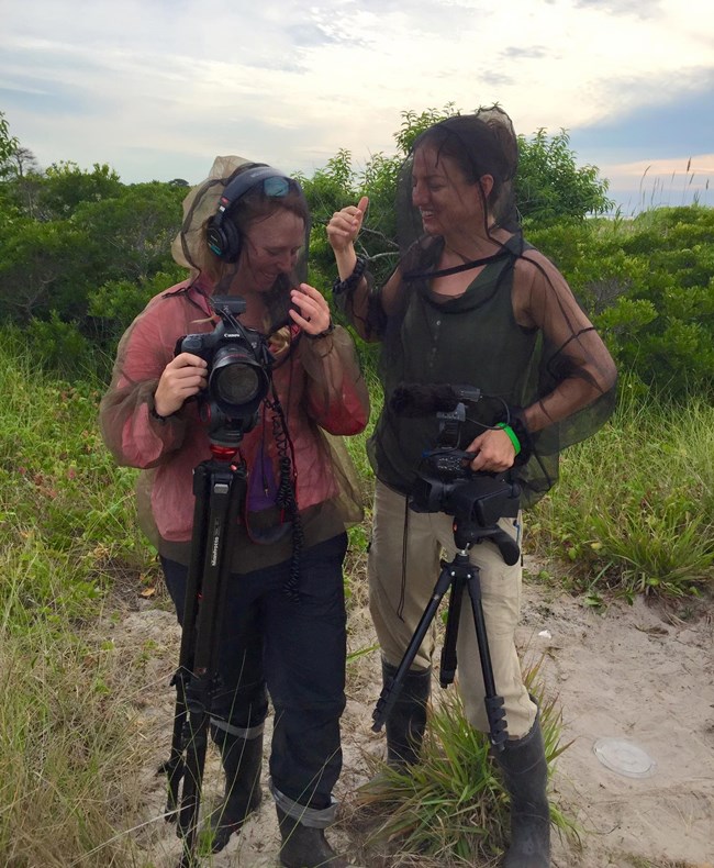 Two women stand in a marsh with camera eqipment