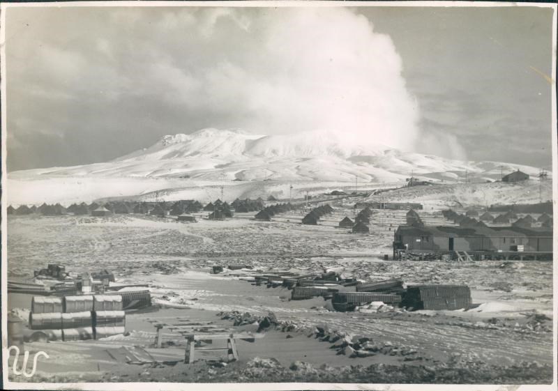 Black and white photo of small tents and buildings with snowy mountain in background.