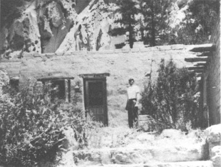 A black and white photo of a man stands in front of an adobe house.