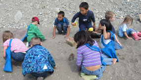 a group of children on a light colored sandy beach. The children are digging through the sand, attempting to find symbolic archaeological items.