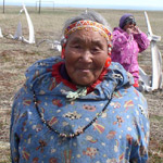 A native elder in Chukotka Russia. She is wearing a traditional shirt and headband. Whale bones are standing on end in the background.