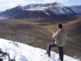 A man standing in snow on a late autumn day on the side of a mountain in Russia. He is overlooking a valley across to a mounting range on the other side.