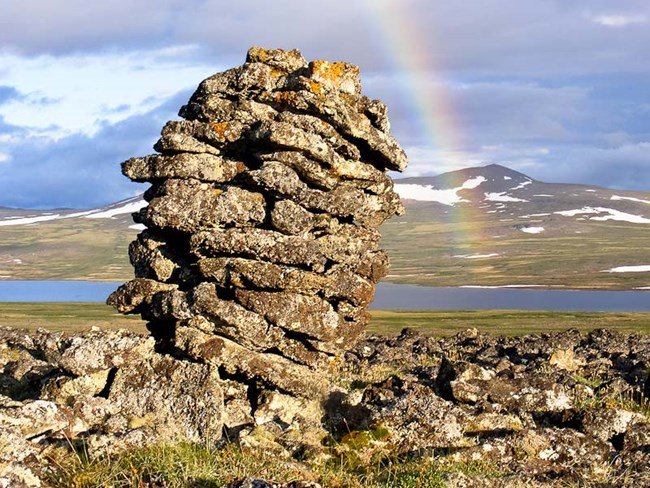 A rock cairn standing above the tundra with a rainbow arching across the background.
