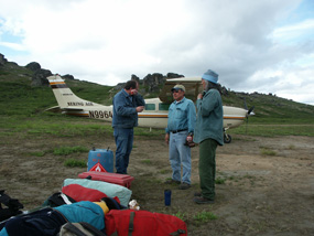 Three men standing next to their outdoor gear with a small fixed wing aircraft in the background.
