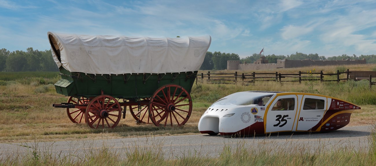 A sleek solar car rests next to a covered wagon with an adobe fort in the backgrouns