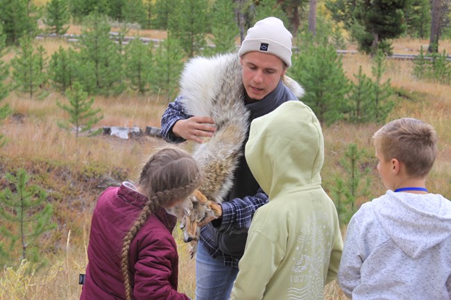 A man facing the camera holds a light grey coyote pelt.  Several chidren are looking at the pelt, and one child is touching the pelt.
