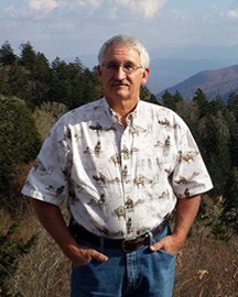 Older male with a mustache standing in a field of grass and trees with a mountain in the background.