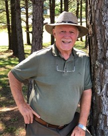 Older male with a cowboard style hat wearing a green shirt, leaning up against a tree.