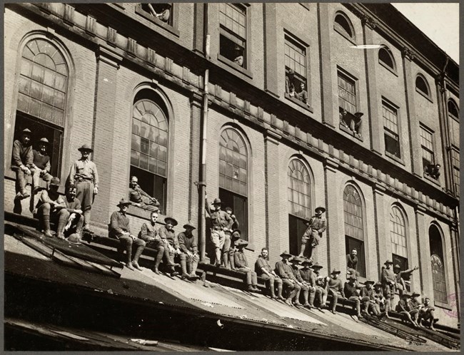 uniformed men on the awning of a building. some are seated while others are standing.