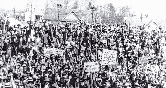 a large group of people holding signs and waving to the camera with houses in the background