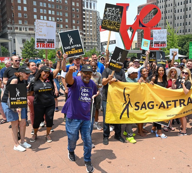 a group of people holding picket signs and a large yellow banner that says SAG-AFTRA