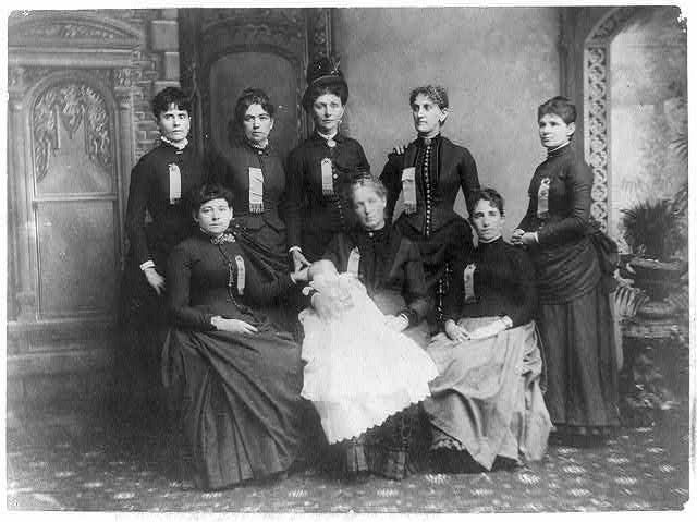 8 women and one infant in dresses, seated for the camera.