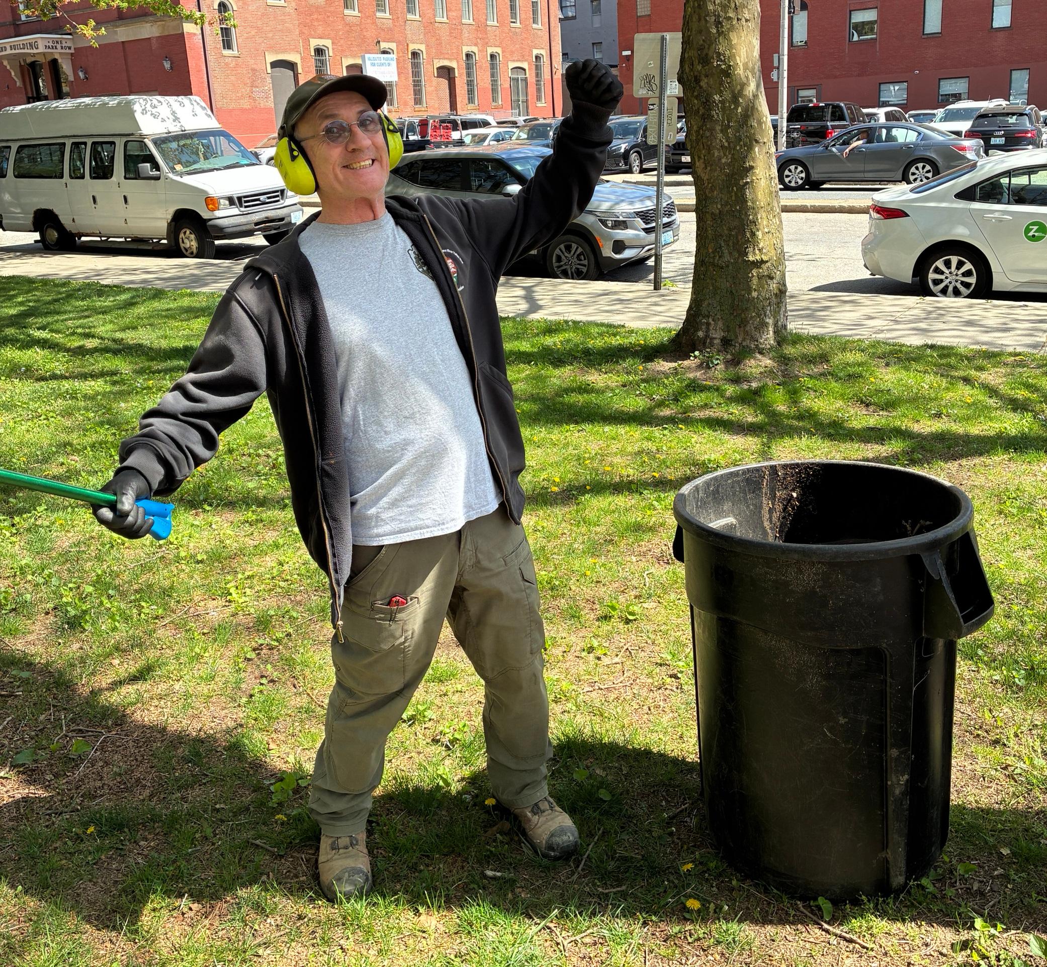 Man in black sweatshirt and wearing hat stands next to trash bucket with hands raised and a picker in one hand