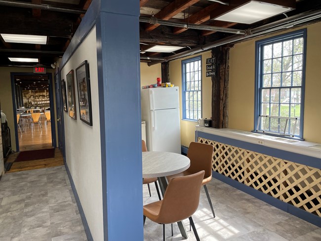 Wall dividing a hallway with pictures on the wall and sitting area with tables, chairs, and refrigerator