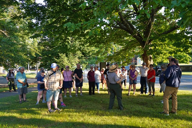 Group of people stand around ranger. All standing underneath a tree