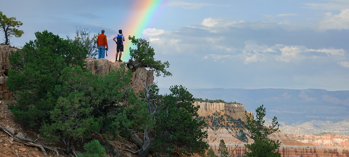 two people stand at the edge of a cliff watching a rainbow