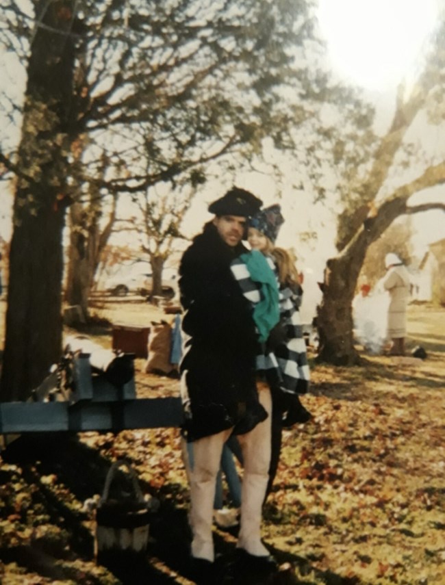 Man dressed in Revolutionary War attire holds young child outside as they pose for a picture.