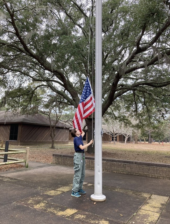 Person lowering the U.S. Flag at flagpole. Wooden building and large tree are seen in background.