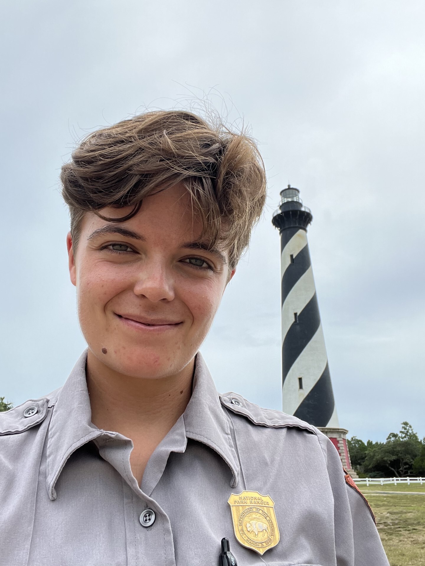 Ranger smiles at the camera with Cape Hatteras Lighthouse in background. Lighthouse has black and white diagonal stripes.