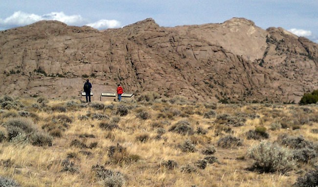 Two people stand in front of an outdoor exhibit while looking at a large rock formation called Split Rock in Wyoming with sagebrush.