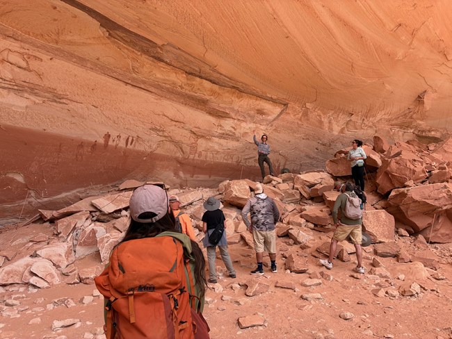 A person wearing an orange backpack looks up towards a group and a ranger presenting in front of a wall with pictographs