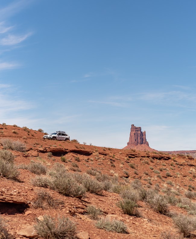 A white Jeep drives along a backcountry road with canyon towers behind it