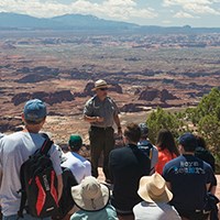a group of people and a ranger at an overlook with canyons in the background