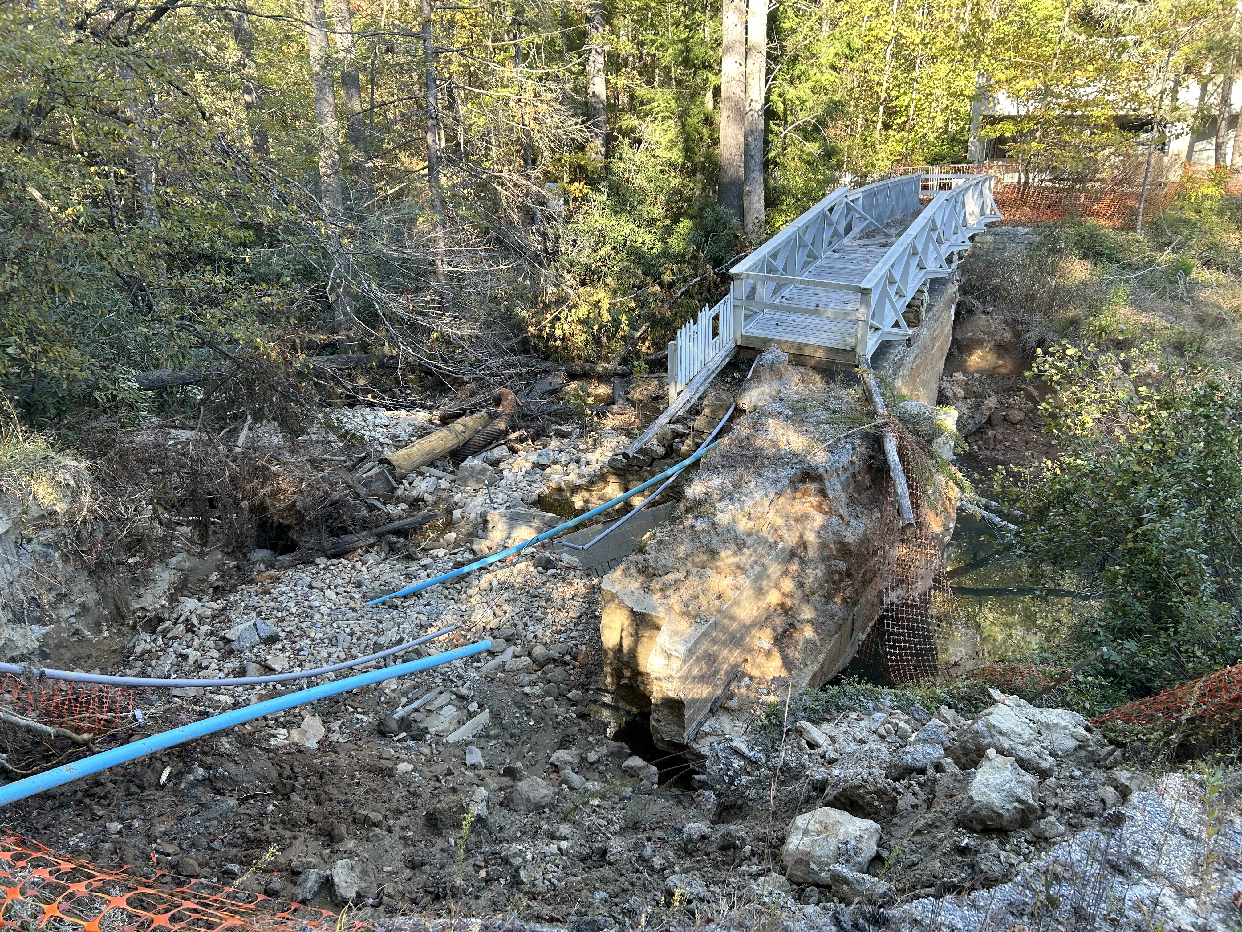 A boardwalk and thick concrete wall end abruptly. They are surrounded by broken pipes and debris. Orange safety fencing is visible in the distance and foreground.