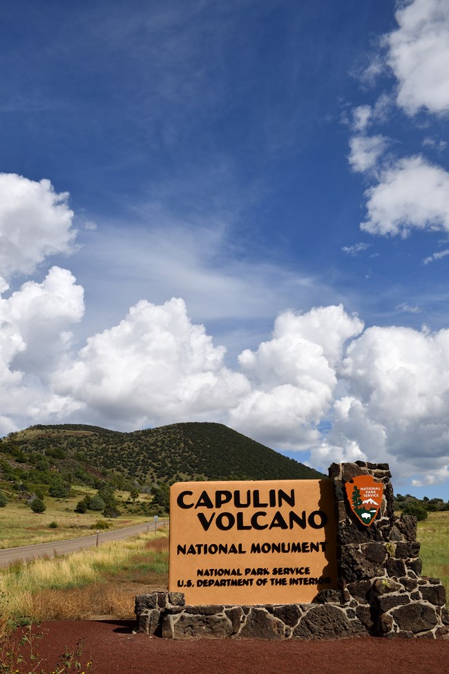 An entrance made of lava rock has states "Capulin Volcano National Monument, National Park Service, Department of the Interior".  The sign also includes the National Park Service Arrowhead Logo.  The sign sits in the foreground of a forested volcano.
