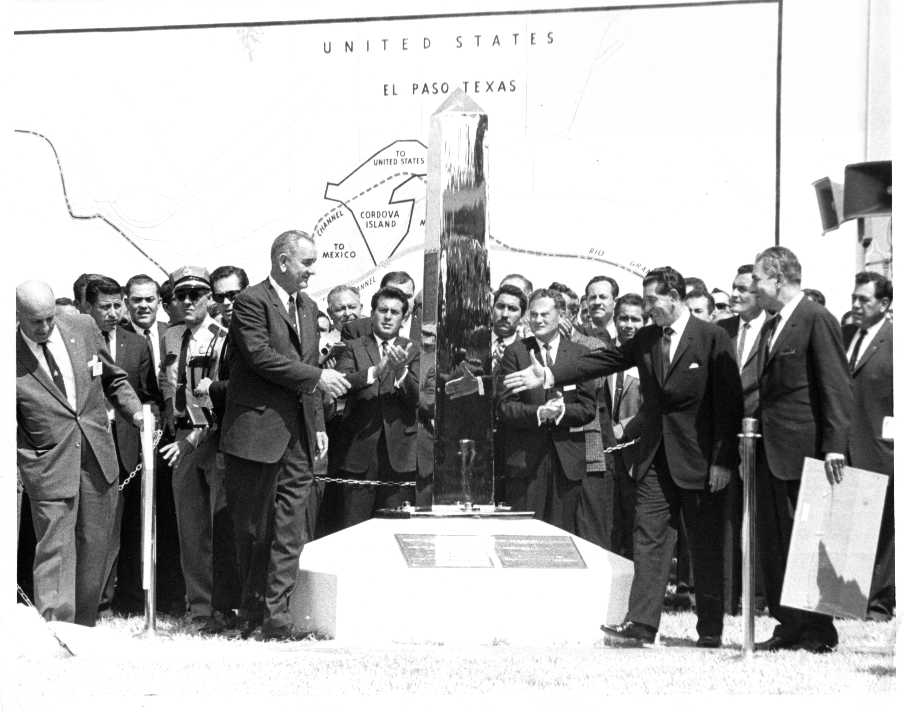 black and white, two men reach to shake hands in front of reflective obelisk and a crowd of people
