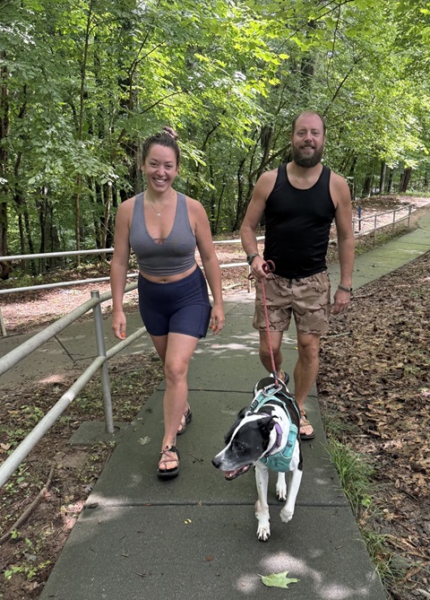 Two smiling adults walk a dog on a leash in the woods.