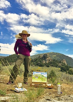 The artist stands with her binoculars next to her painting easel with a painting depicting the City of Rocks scene in the background.