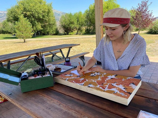 Young woman with painting supplies working on a painting seated at a picnic table in a green park setting.