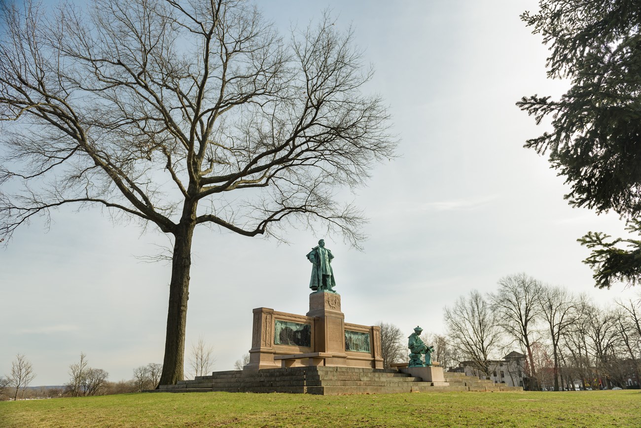 A memorial statue with two figures set upon a hill underneath a tree on a sunny day.