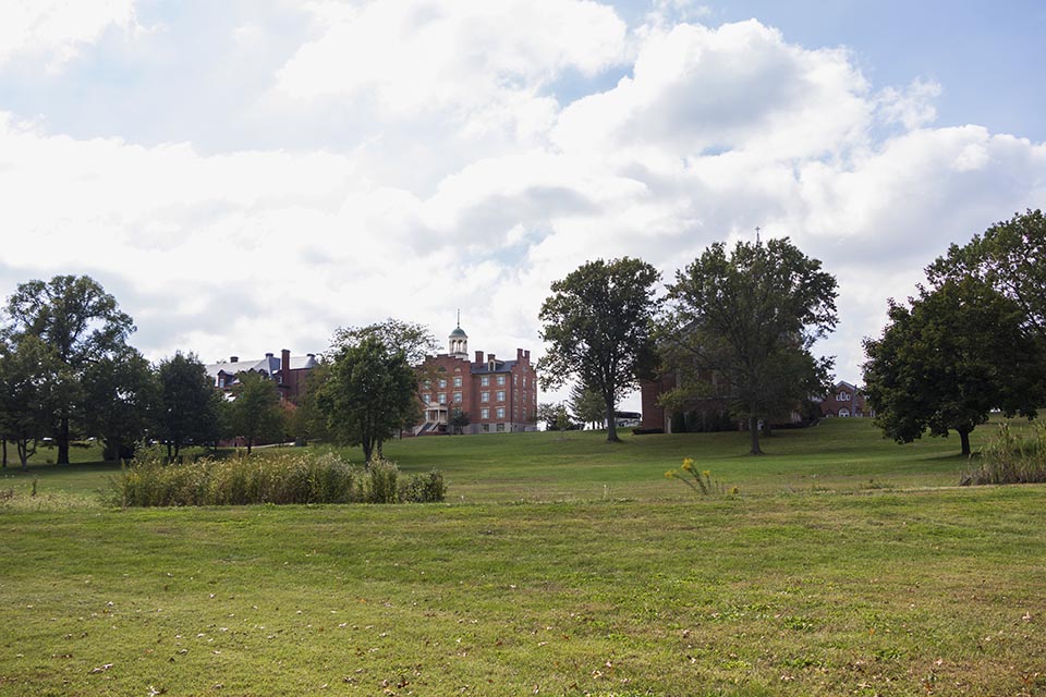 A young man sits on a white fence looking up the hill at a large building with a white cupola.