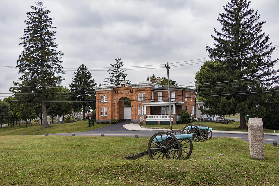 A black and white picture is superimposed on a modern picture of a red brick building shaped like an arch with two cannons in front of the structure.