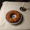 A bundt cake on a tablecloth. Next to it, a single lit candle. 