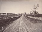 An 1800s photograph shows a roughly paved road lined with stone walls passing through farmland. 